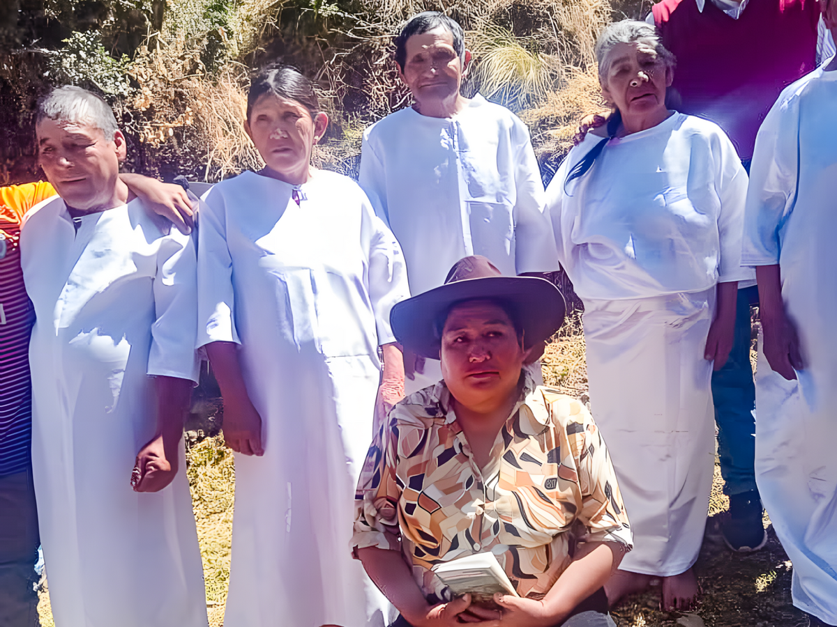 Eine peruanische Ketschua-Frau mit Hut kniet mit Blick in die Kamera vor einer Gruppe älterer Menschen, die als Täuflinge in weißer Kleidung in einer Reihe hinter ihr stehen.