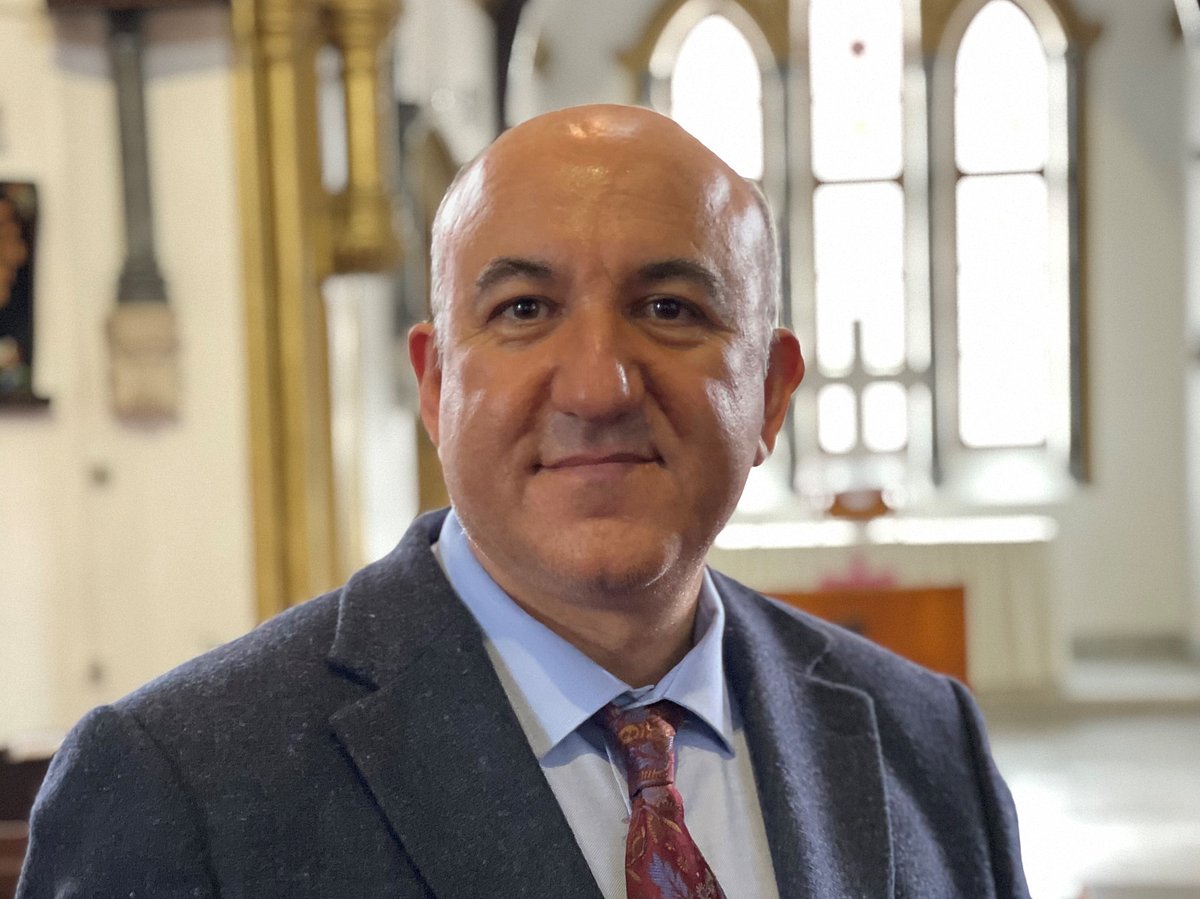 EIn Pastor im Anzug steht in seiner Kirche, im Hintergrund sind Kirchenfenster zu sehen.