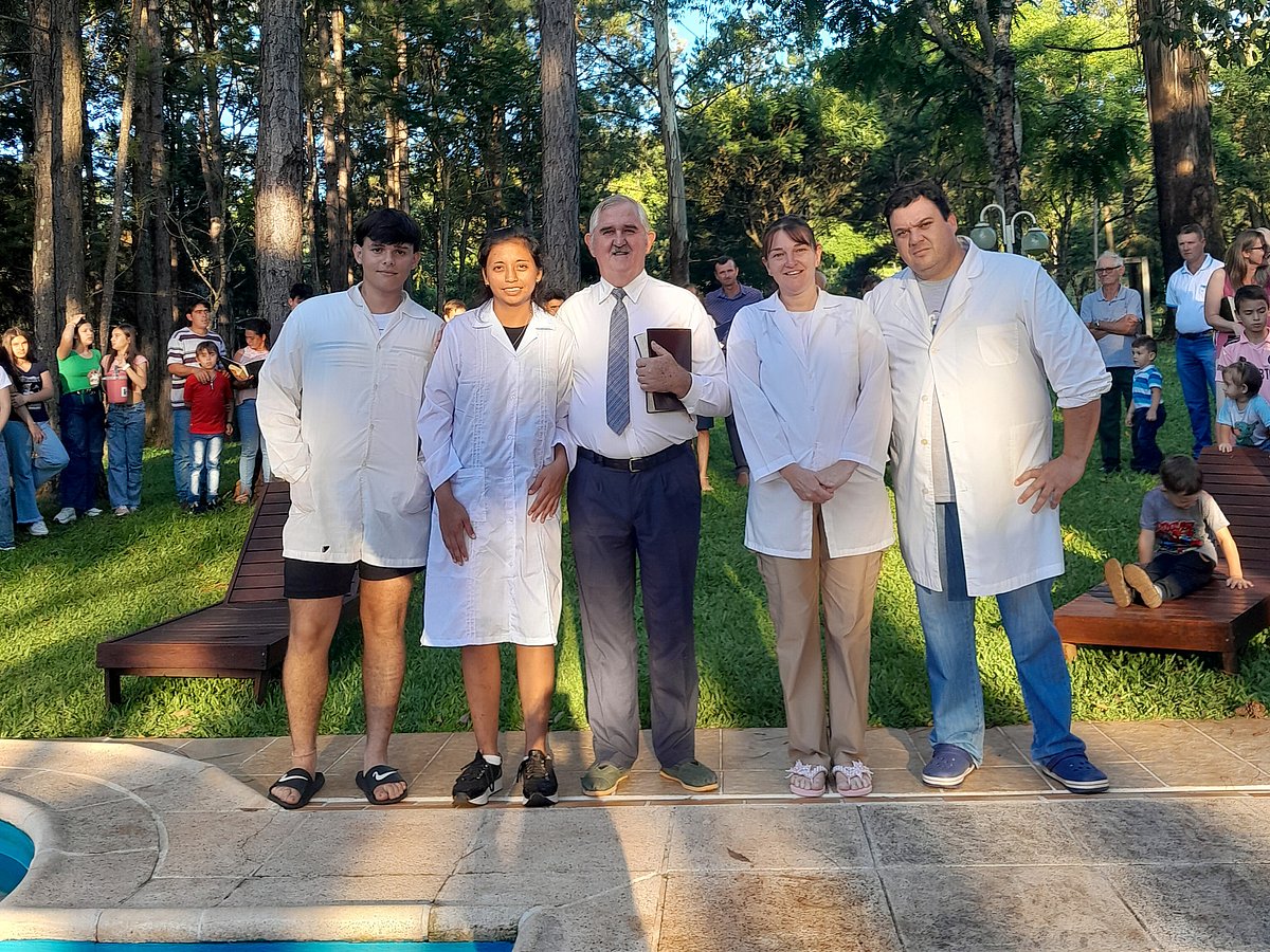 Eine Gruppe von fünf Erwachsenen steht mit weißen Mänteln gekleidet am Rand eines Schwimmbeckens und wartet auf die Taufe