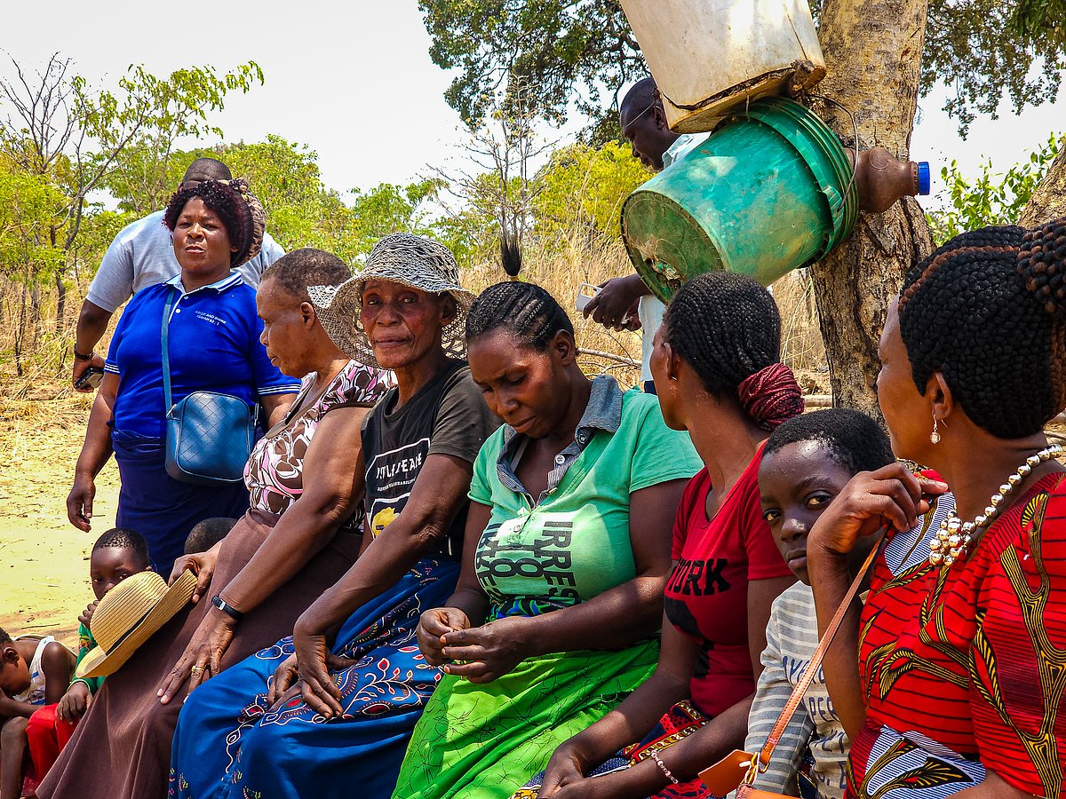 Gruppe von zehn Menschen, überwiegend Frauen, sitzen und stehen unter einem Baum in ländlicher Umgebung in Sambia