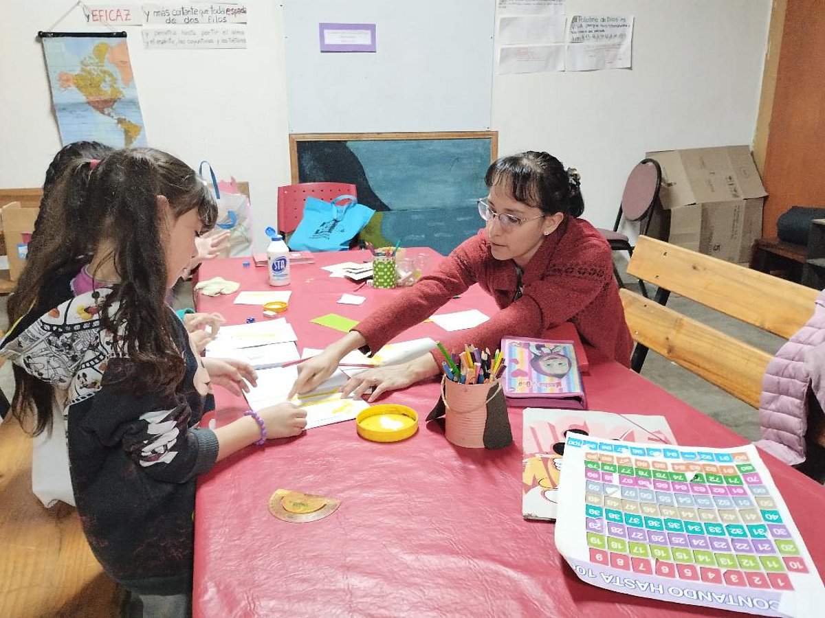 Kinder sitzen an einem Tisch und erhalten von einer Frau Hilfe bei den Schularbeiten