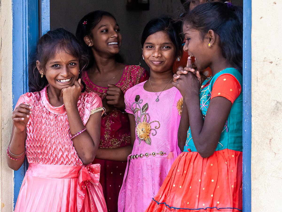 Mehrere indische junge Mädchen stehen in rosa und bunten Kleidern in einem Türrahmen und lächeln in die Kamera