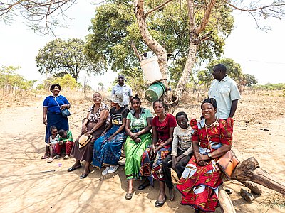 Zehn Personen, darunter Frauen und Kinder, sitzen und stehen unter einem Baum in einer ländlichen Umgebung in Sambia.