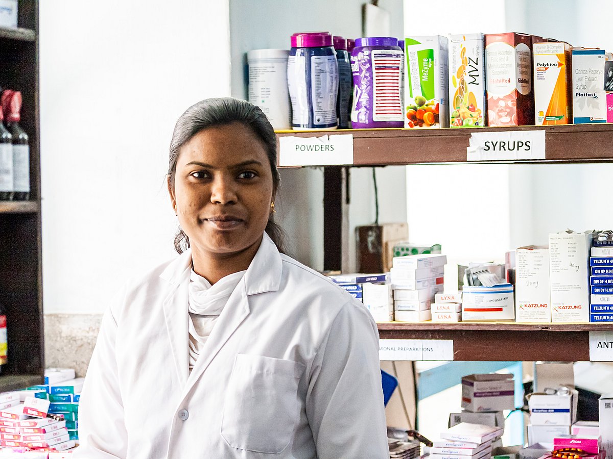An Indian woman in a white coat stands in front of a shelf with medicines.