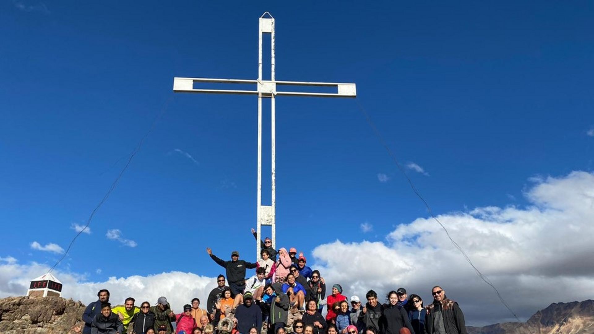 A group of people is standing on the top of a mountain, behind them is a big white cross and blue sky