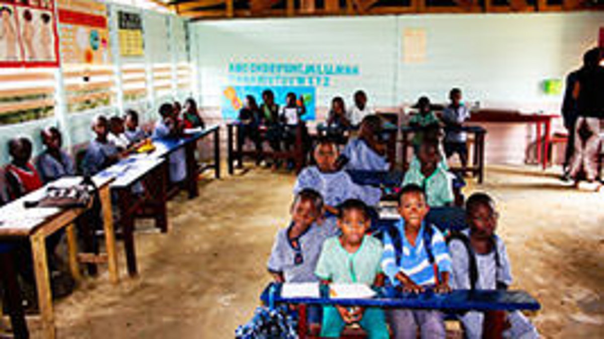 Schulkinder sitzen in einem Klassenzimmer in Sierra Leone mit Holztischen und -bänken, einige tragen Schuluniformen, im Hintergrund eine Tafel mit Text