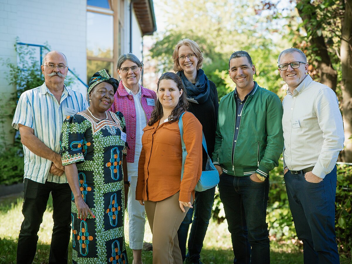 Eine Gruppe internationaler Menschen steht in einem Garten und lacht in die Kamera.