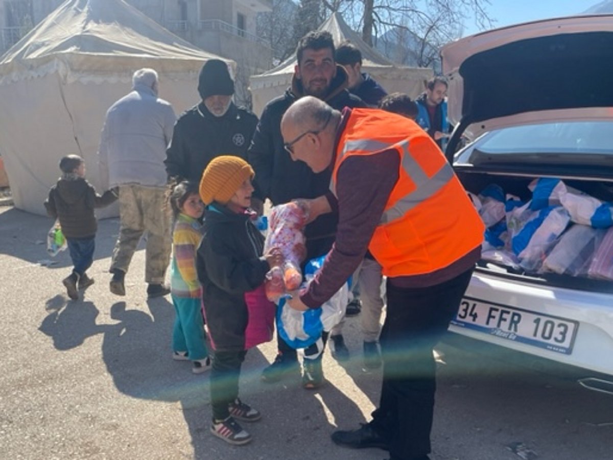 Ein Mann in einer orangenen Warnweste steht vor einem Auto mit offenem Kofferraum und gibt mehreren Kindern Hilfsgüter.